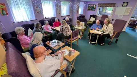 Martin Giles/BBC A group of elderly day centre attendees are sitting in chairs in a semi-circle enjoying crafts and games.
