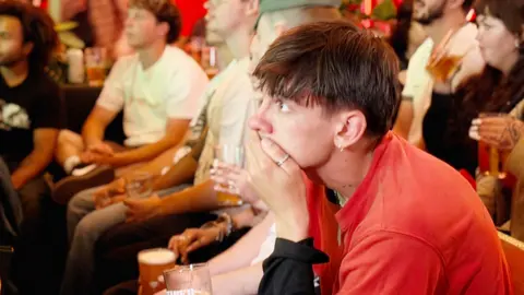 Joey, a young man in a red top, watches on nervously as England play Croatia. He has dark hair and holds a pint glass in his right hand