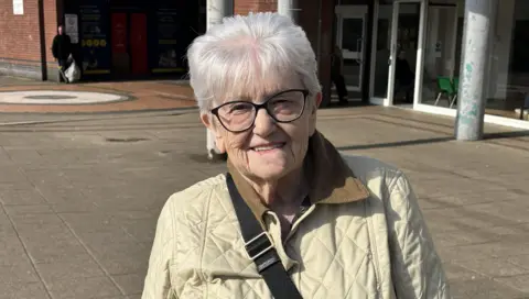 Anne is a smiley older lady with short, well-styled white hair and large fashionable glasses. She wears a beige anorak. A row of shops runs behind her.