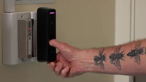 A man's arm with three bee tattoo uses his thumb on a biometric scanner 