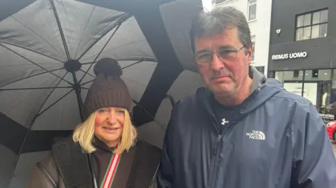 Anne and Peter on the high street. Anne holds an umbrella and is wearing a brown coat, a brown hat and has short blonde hair. Peter is wearing a blue jacket and is wearing glasses, he has short dark hair with grey strands. 