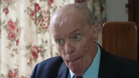 A picture of an elderly man wearing a light blue shirt and dark suit jacket. He is sat in front of flowery curtains in his home