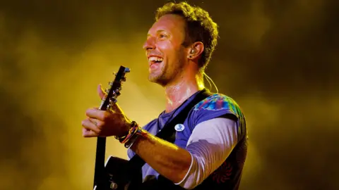 Chris Martin smiling while performing on stage and holding a guitar. He has short dark hair and is wearing a multicoloured T-shirt with long arms. He is smiling and behind him is dry ice smoke lit by a yellow light.
