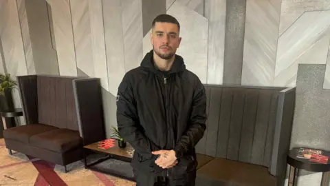 A young man. His arms are folded across his body. He is wearing all black clothing and has short hair. 