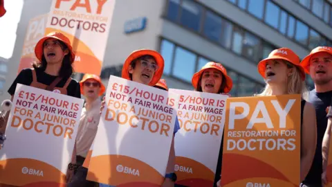 A group of junior (now called resident) doctors holding placards outside a hospital in London last year. They are holding signs saying "£15 an hour is not a fair wage for a junior doctor" and wearing orange hats with the BMA logo on them. They look like they are singing or chanting.