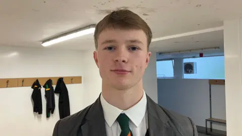 Pupil Michael. He has light brown hair, wearing a grey blazer, white shirt and green tie. He is standing in a sports changing room, behind him black tracksuit items are hanging on pegs.