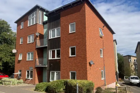 Owen Sennitt/BBC A block of flats with red bricks and a balcony can be seen, with shrubbery in front of them.