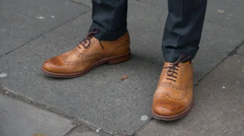 A close-up of a pair of brown brogue shoes by Grenson, on a man's feet, standing on a road. They are laced up and the man is wearing blue trousers.