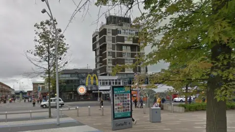 Google 1980s eight-storey office block in red brick with white lines around each floor and white window frames. The bottom floor is occupied by shops and there is a large letter M in yellow to the right of the eight storeys. There is a small tree to the right of the building and a larger tree in the foreground to the left. There is a road passing the office building and a dynamic advertising board is visible in the centre of the picture.