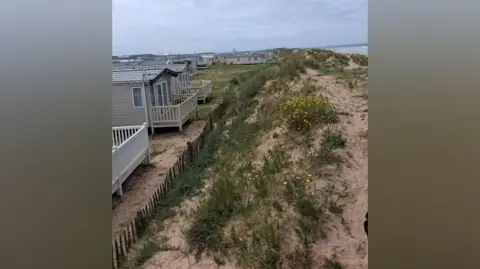 Carl Quartermain The sand dunes are dotted with green shrubs, some of them flowering with yellow buds. A short wooden fence separates the dunes from the static caravans on the shore. The ocean is on the right, in the background.