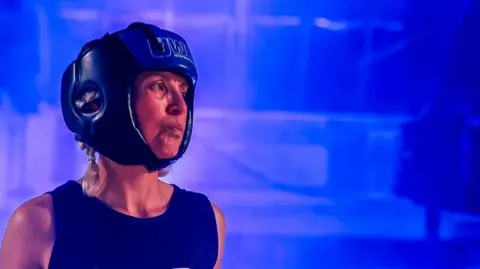 Teresa Colman Teresa wearing a blue sleeveless boxing jersey and a blue helmet standing inside the boxing ring with blue lights. 
