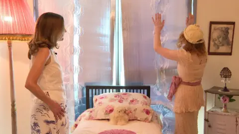 Flora shows Leyla the heat reflective panelling on the windows in the bedroom 