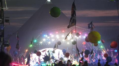 Photo of the pyramid stage at Glastonbury Festival 2024, when Coldplay headlined. A large crowd can be seen at the bottom of the frame, and balloons and flags are in the air. The stage is lit with different coloured lights.