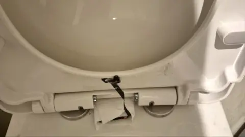 The lifted lid of a white toilet seat, with a black camera tucked under its rim and a wire leading to another part of it wrapped in kitchen roll