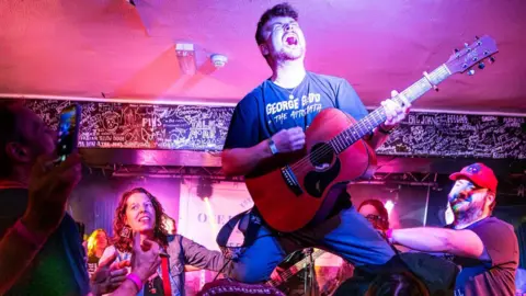 Harrison Rimmer, singing emotively while playing the guitar, knelt on top of a human pyramid in The Fulford Arms, York.