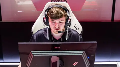 A young man wearing a look of intense concentrations sits in front of a black computer monitor. He's wearing a large, expensive-looking gaming headset with a microphone attachment, and sitting in a large, white gaming chair.