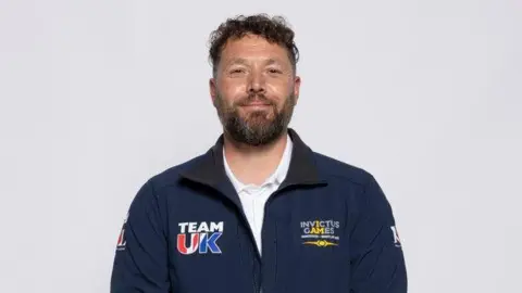 Royal British Legion Daniel Bennet is wearing a navy blue Invictus Games Team UK zip jacket. He has his arms down by his side and is smiling at the camera. 