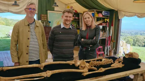 BBC/Rare TV Oscar Aldred and Benjamin Neil showing Alice Roberts the remains of a man with trepanning injuries in the Digging for Britain tent. In front of them is a trestle table and resting on a black sheet is the skeleton of a man with its head at the top right. The tent doors are open and there are views of the countryside beyond. 