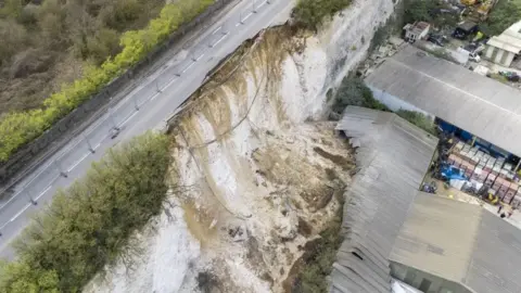 High Profile Aerial  A road along a chalk clifftop with industrial buildings underneath. The road has partly fallen away after a landslip.