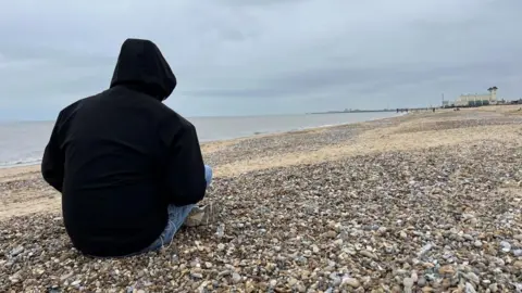 Andrew Turner/BBC A picture of Leon in a black hoodie, with the hood covering his face to protect his identity. He is pictured on Great Yarmouth's central beach, with the Wellington Pier visible in the distance.