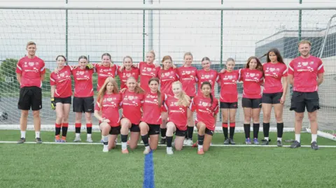 A group of girls in red T-shirts and black shorts in a line, with five girls crouched in front. A male coach is either side of them, with a football goal behind.