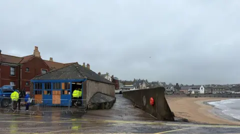 A old boathouse is being worked on by people in high vis jackets and to the right of the picture is a slipway that lets you see right along to beach. the coast line of Eyemouth is visible.