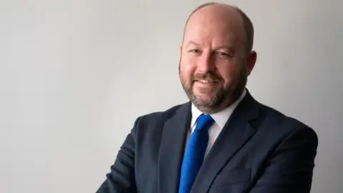Contributed Nick Timothy is smiling at the camera and is wearing a dark suit jacket over a white shirt with a bright blue tie. He is posed against a plain light-colored background with arms crossed. 