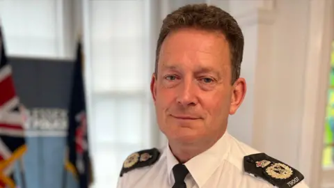 Ben-Julian Harrington stands in a white shirt with black epaulettes and a black tie. He is standing in front of a lectern with an Essex Police banner in the background.