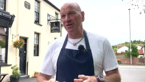 A restaurant owner wearing a navy blue apron and white t-shirt stands outside his restaurant while explaining the impact traffic will have on his business