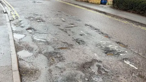 Mark Slack A residential road with lots of small potholes, many of which are filled with water. There are double yellow lines on either side of the road.