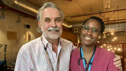 Ashley Brown is seen in a pink and white striped shirt with a rainbow NHS lanyard stands next to Mainga Bhima in burgundy medical scrubs with a stethoscope around her neck. They are both smiling in an indoor setting with industrial-style lighting in the background.