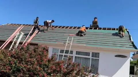 Facebook A picture of four men working on the roof of a bungalow on a sunny day. You can see the wooden structure of the roof at the very top. The rest is covered in a green blue fabric and has red strips of wood going along it length ways.