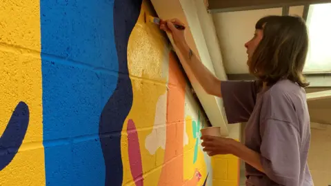 sarah hoyle Sarah paints a colourful image onto a white brick wall. She has brown hair with a fringe. Her hair comes down to the top of her shoulders and ends with a flick.