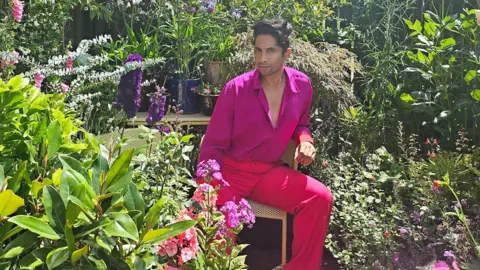 Kamal Kaan A man in pink trousers and shirt sits in his garden surrounded by flowers