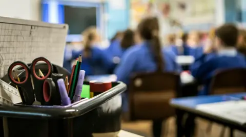 PA Media In the blurred background of the photo are a group of children in blue uniforms in a primary school classroom looking at a board. In the foreground of the photo is a clear image of a pot of stationary