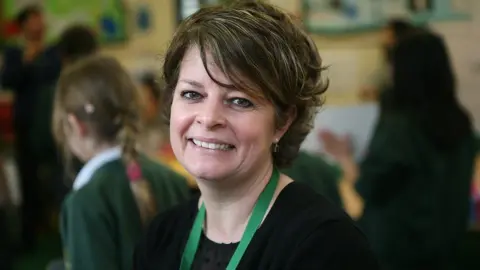 Ruth Perry smiling and wearing a black top with green lanyard. She is in a classroom with blurred children in the background.