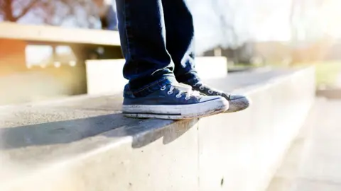 A child's feet, wearing jeans and blue baseball boots, sitting on a pair of concrete steps.