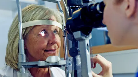 Older woman with shoulder-length blonde hair having a photo taken of her eye by an optician with specialist equipment. A light is shining in one of her eyes, lighting up her pupil.