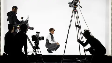 Daniel Boud Cynthia Erivo dressed in a suit on stage against a white backdrop with three cameras pointing at her. 