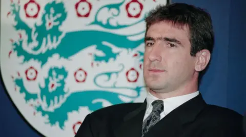 Getty Images Dark haired man in dark suit sits back on a chair with an England football crest in the background