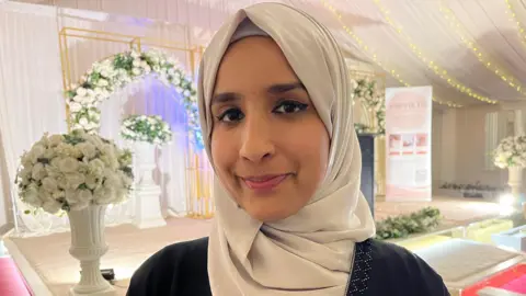 Nicola Haseler/BBC Manisha smiles at the camera, wearing a white silky headscarf and black top. There are wedding-style flower arrangements behind her, and a flower arch.