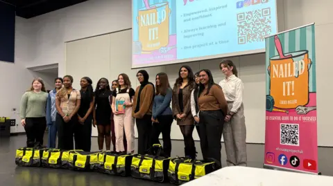 Ben Schofield/BBC A line up of people standing inside and looking off camera to the left. In the middle of the line is Laura Deller, who is wearing pink dungarees and a black top. She is holding a copy of the book Nailed It!, which she has written. Either side of her are female sixth form students from Bedford Academy school, six on either side. They are not wearing a school uniform, but are smartly dressed in their own clothes. In front of the line up are several heavy-duty looking DIY carryalls. A vertical banner promoting Nailed It! is on the right of the image. It has a cartoon picture of a pot of paint, with a brush dipped in, a spanner and a tape measure on it. The caption reads "The essential guide to do it yourself".