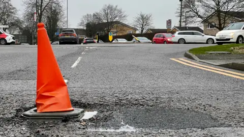 An orange traffic cone placed inside a pothole with a busy mini-roundabout in the background.