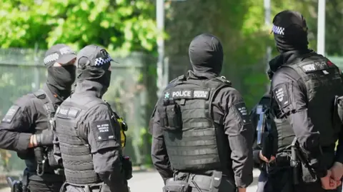 A group of armed police officers wearing baseball caps and face coverings. They are also wearing protective flak jackets. In the distance are trees.