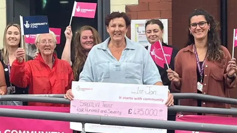 Members of the Streetlife charity smile and hold up National Lottery community fund pink flags and a cheque for £475,000 outside the entrance to Streetlife's base.