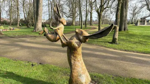 A bronze statue of the two hares from the Guess How Much I Love You book in Lisburn Castle Gardens on a sunny day. The large hare is holding a much smaller hare in the air with its paws. There are tall trees, lawn and pathways in the background.