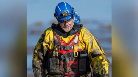 A coastguard crew in specialist uniform covered in mud.