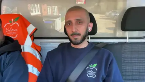 Raaj sits in one of his vans and looks to camera with a serious face