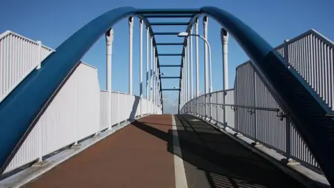 Bob Jones/Geograph The Jane Coston Cycle Bridge over the A14 between north Cambridge and Milton. The cycle and pedestrian bridge has white railings and is arching up into blue sky.