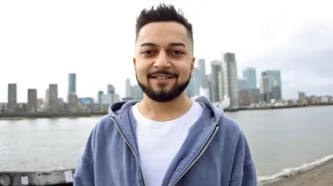 cicelygrace Kishan standing outside wearing a white top and blue hoodie, he is standing with high rise buildings behind him. He is looking straight at the camera and smiling. 